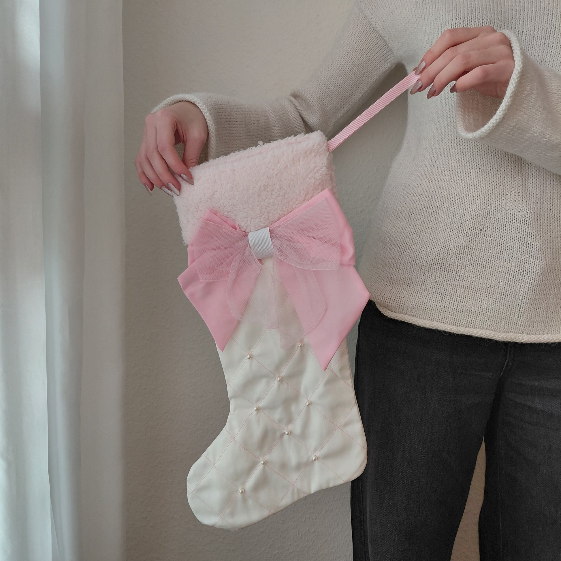 White Christmas stocking with a pink bow held by a person wearing a beige sweater.