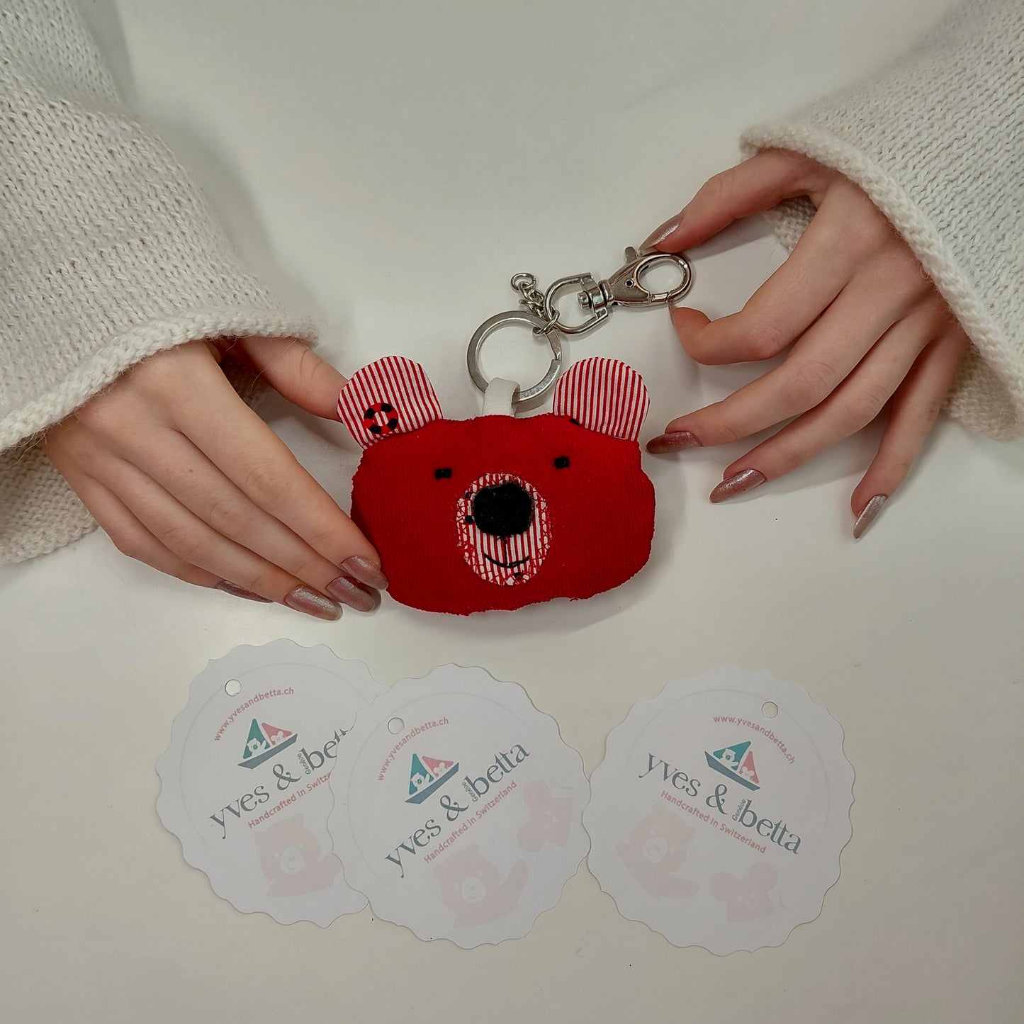 Red bear-shaped keychain held by hands on a white surface with 'yves & betta' labels.