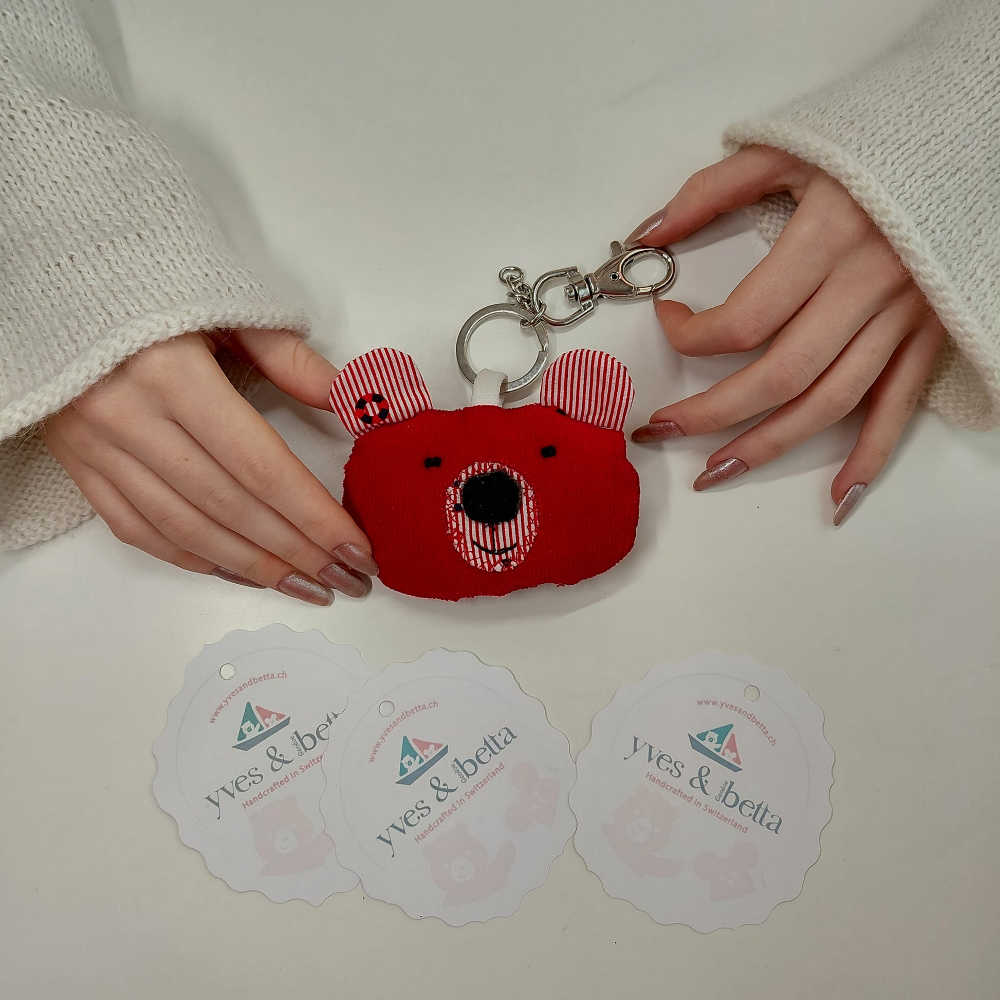 Red bear-shaped keychain held by hands on a white surface with 'yves & betta' labels.