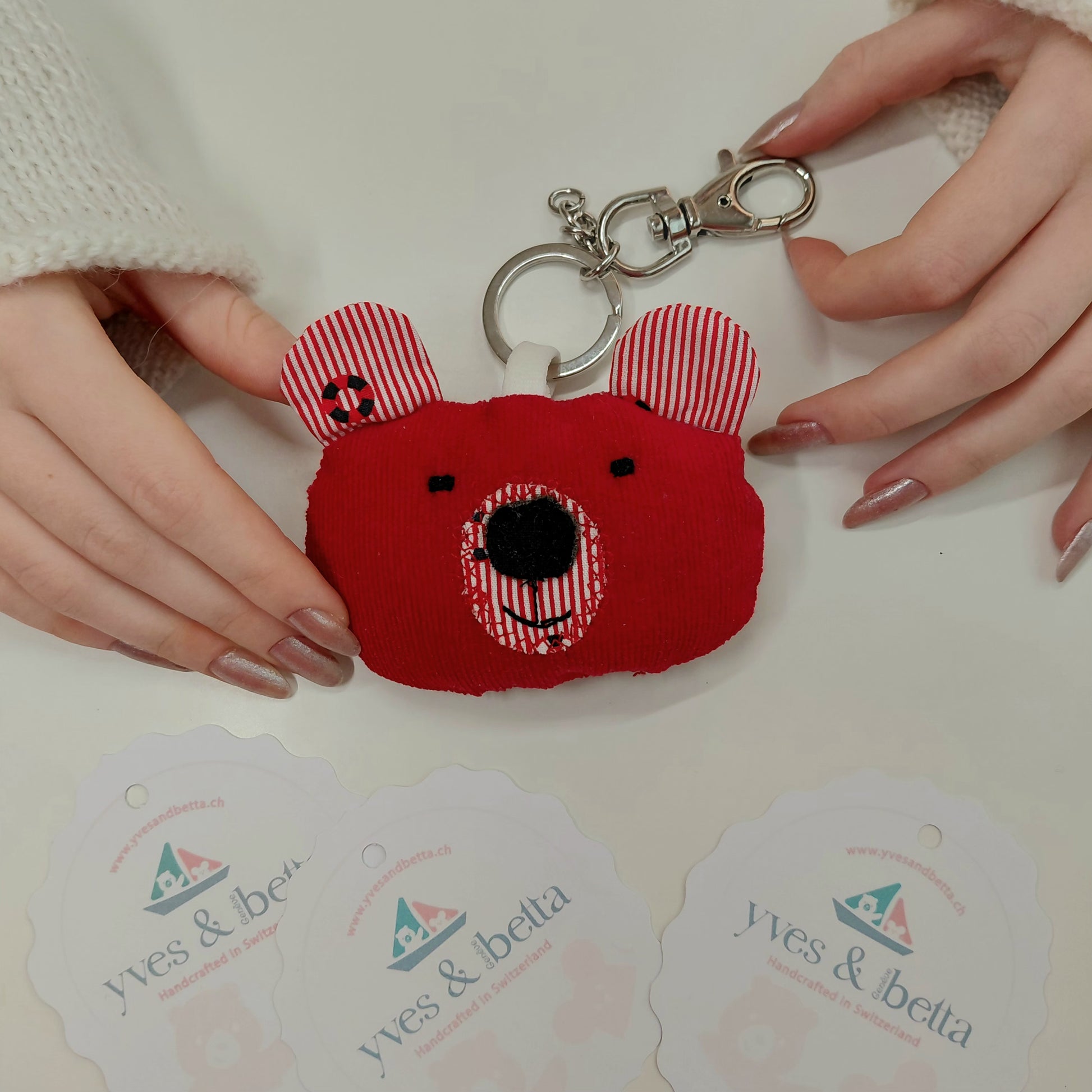 Red plush keychain with striped ears held by hands on a white surface with 'yves & bella' tags.